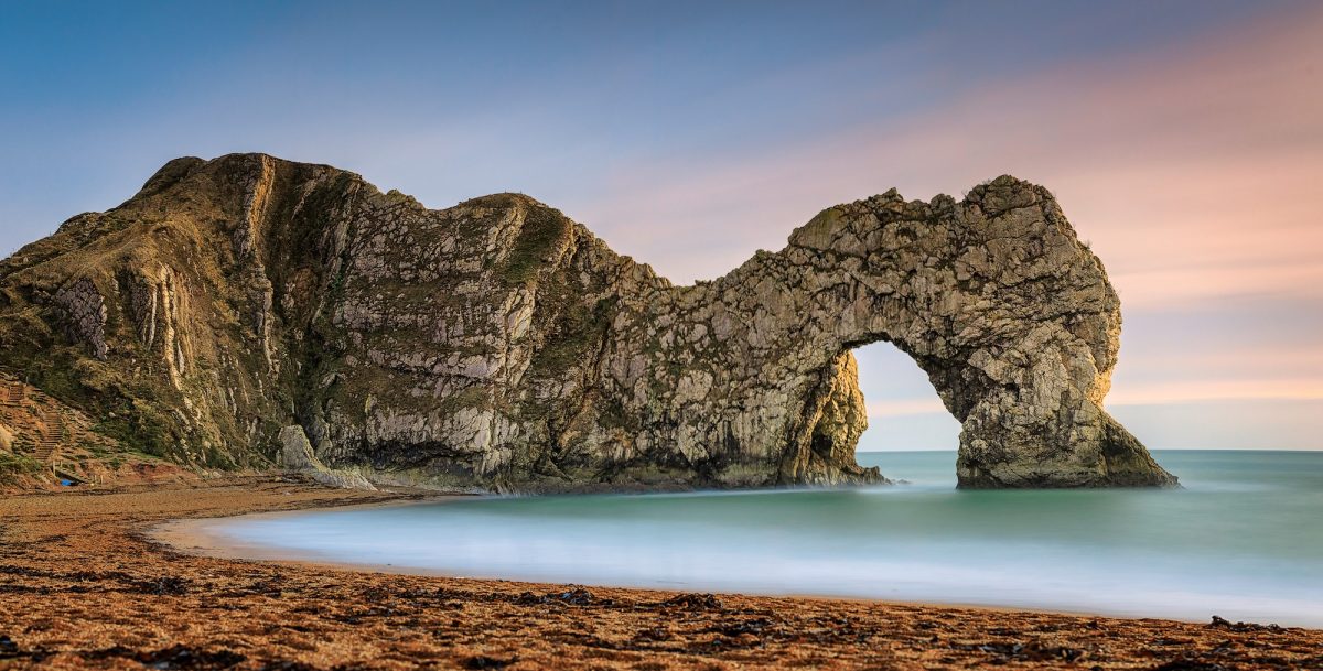 Early morning sun behind the limestone arch at Durdle Door, Dorset, flat sea, sandy shingle beach