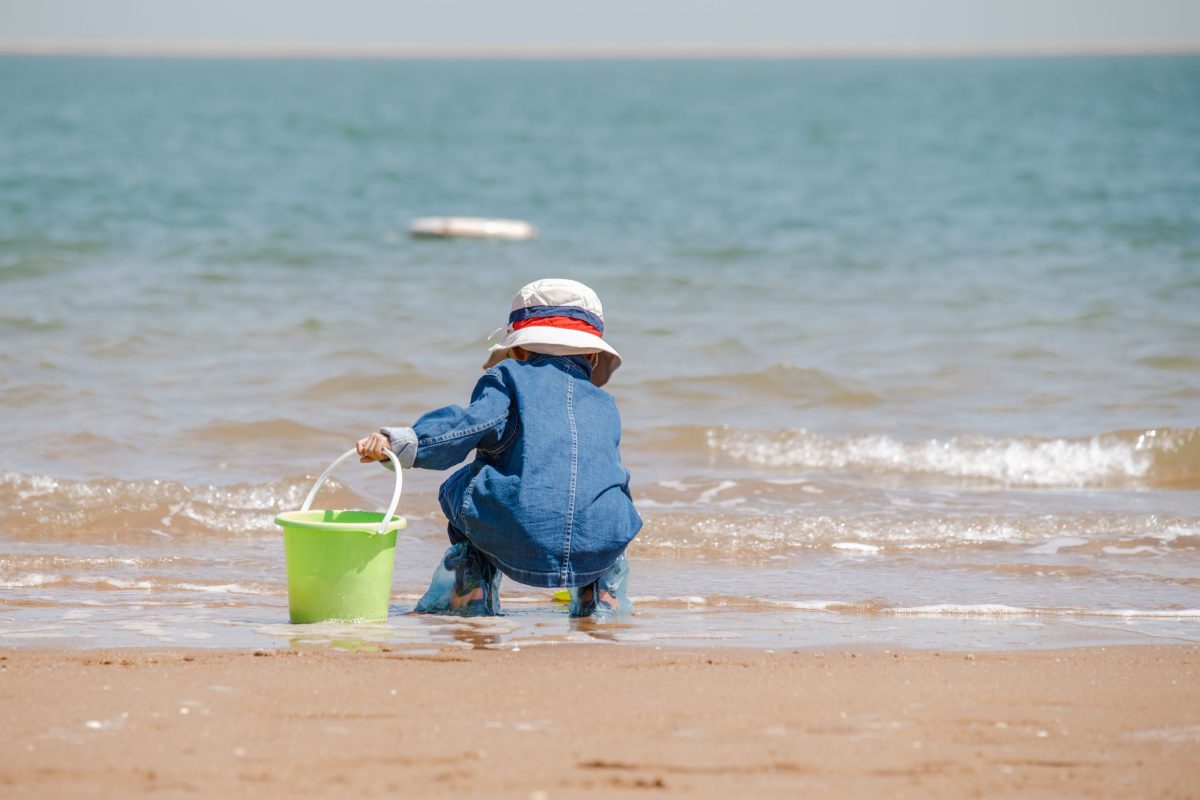 Child wearing a sun hat carrying a bucket on a sandy beach.