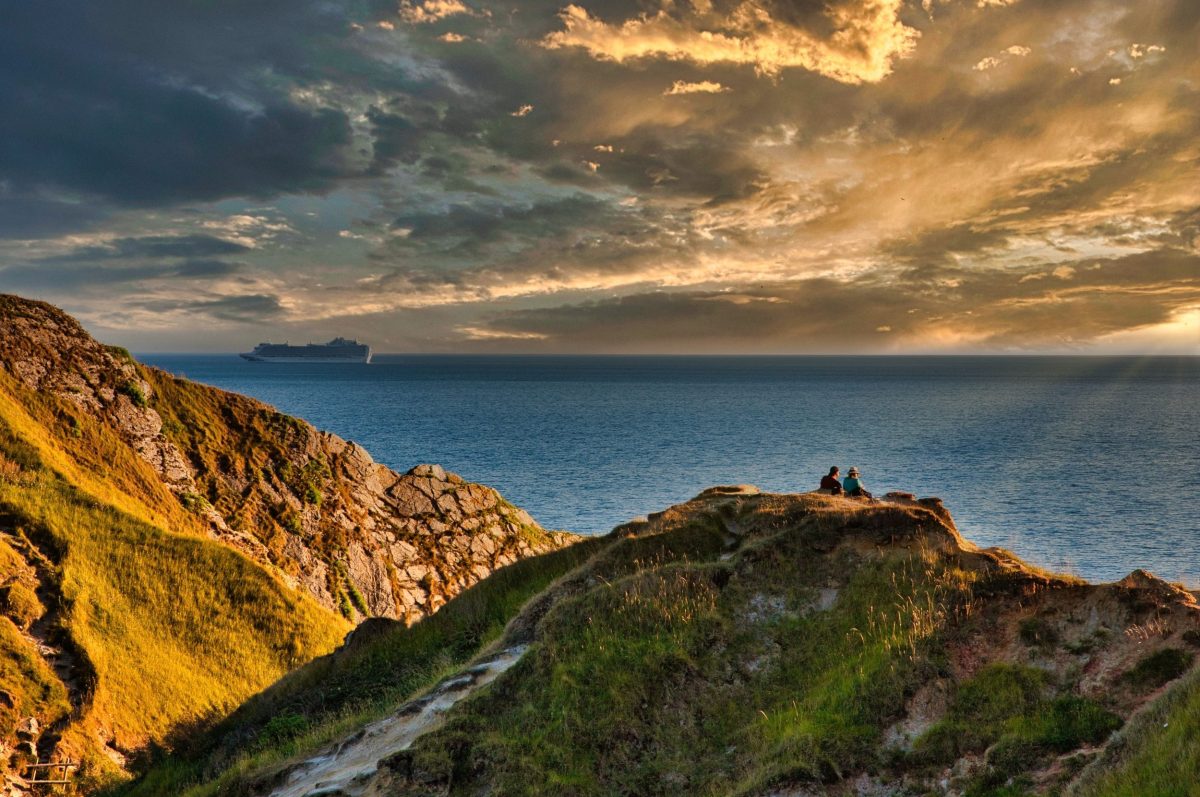 Two people sitting on the cliffs above Lulworth Cove overlooking the sea beneath dramatic skies.