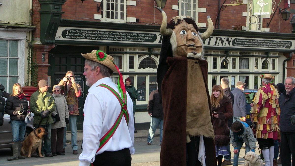 Person dressed as a Dorset Ooser beside a Morris dancer on a busy street in Dorset