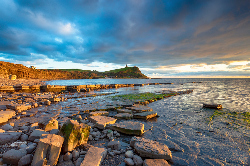Rocky beach at Kimmeridge beneath dramatic grey skies with cliffs in the distance
