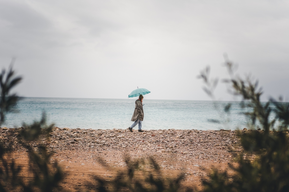 Woman walking with a blue umbrella on a pebble beach in the rain