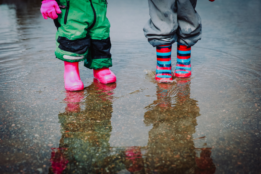 Two children wearing wellington boots splashing in puddles on a rainy day.