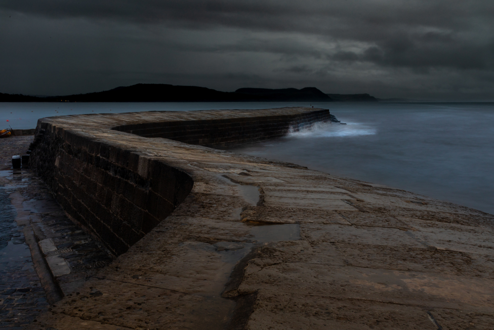 Large waves crashing against the Cobb harbour wall in Lyme Regis beneath dark storm clouds.