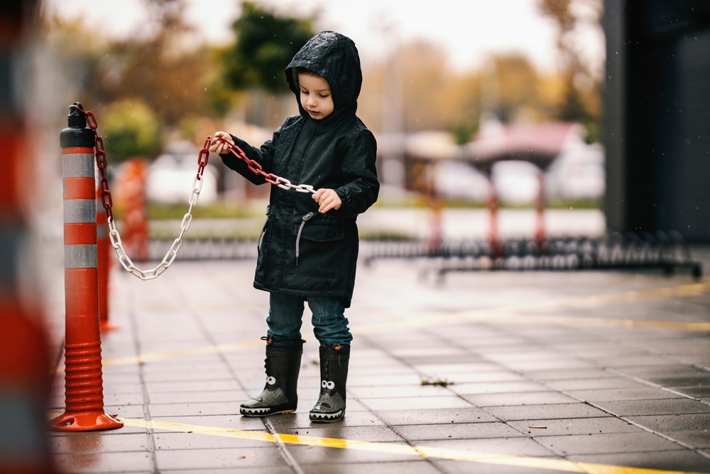 Young boy wearing wellies and a hooded raincoat playing beside a chain on a wet day.